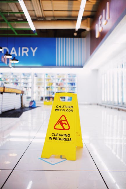 Inside a supermarket aisle with glossy white tiled flooring, illuminated by bright overhead lighting. A yellow caution wet floor sign with black text and a warning icon is prominently placed on the fl