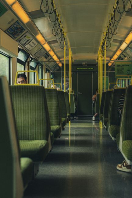 Image of the interior of a bus with green fabric-covered seats arranged in rows along each side of the aisle, which is lined with a non-slip dark grey surface. The bus features yellow handrails overhe